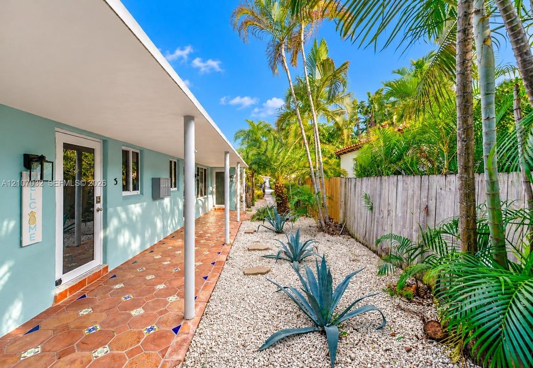 Covered walkway featuring terra cotta tile flooring with decorative inlay, light-blue exterior walls, support columns, and xeriscaped garden beds with stone accents and mature palm trees