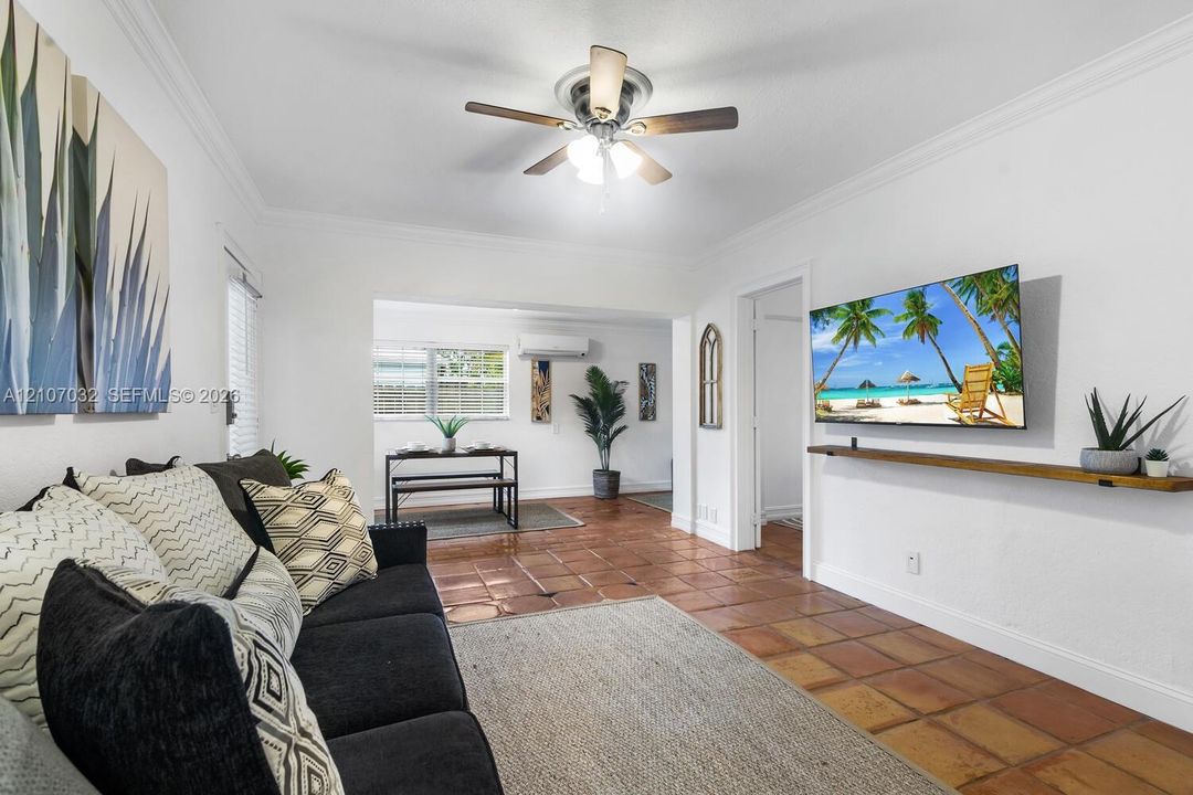 Living space featuring terracotta tile flooring, crown molding, a central ceiling fan with integrated lighting, and a wall-mounted wooden display shelf