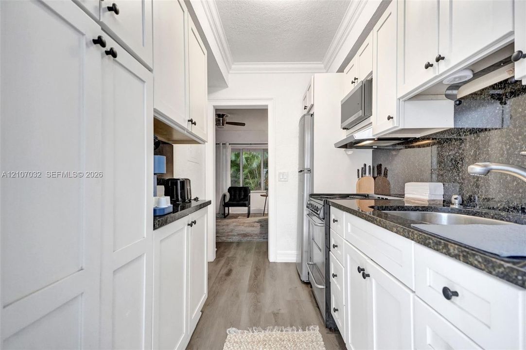 Galley kitchen featuring white shaker-style cabinetry, stone countertops, integrated stainless steel appliances, dark stone backsplash, and wood-finish flooring
