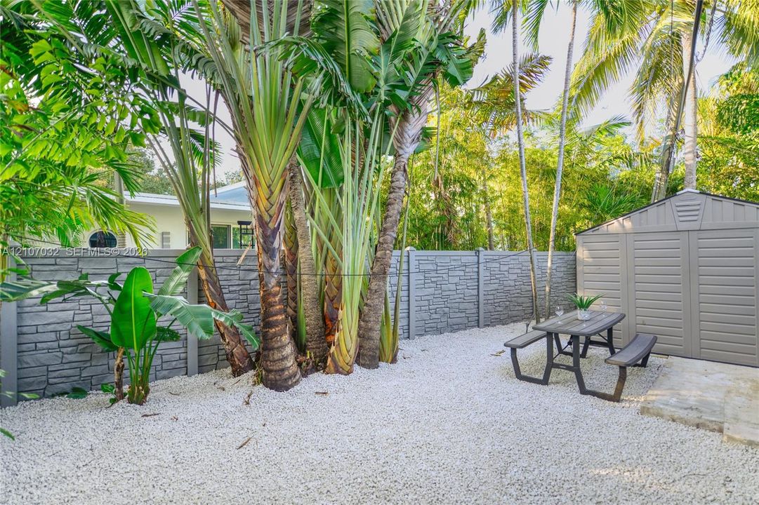 Outdoor space featuring a grey stone-patterned fence, white gravel ground cover, mature palm trees, and a detached storage shed