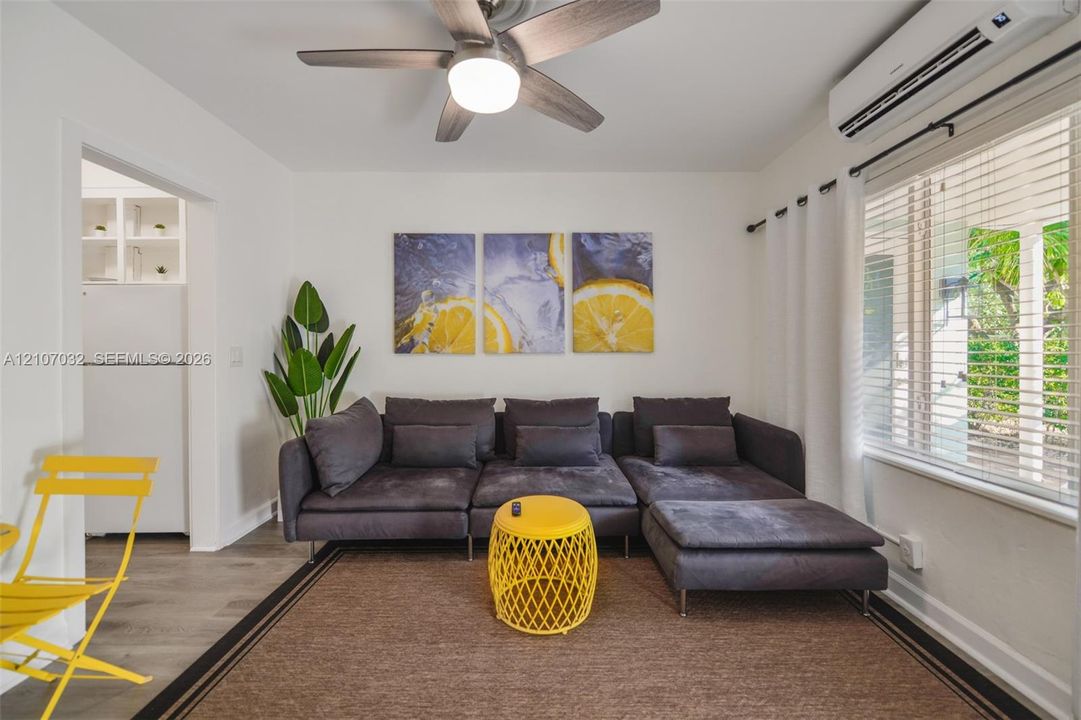 Living area featuring wood-finish flooring, a wall-mounted ductless climate control unit, and a ceiling fan with integrated lighting