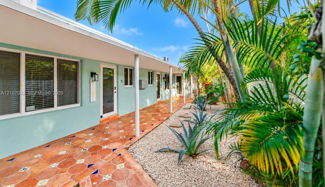 Exterior walkway featuring terracotta tile flooring with decorative inserts, covered porch supported by structural columns, and xeriscaped garden beds with stone ground cover and mature palms