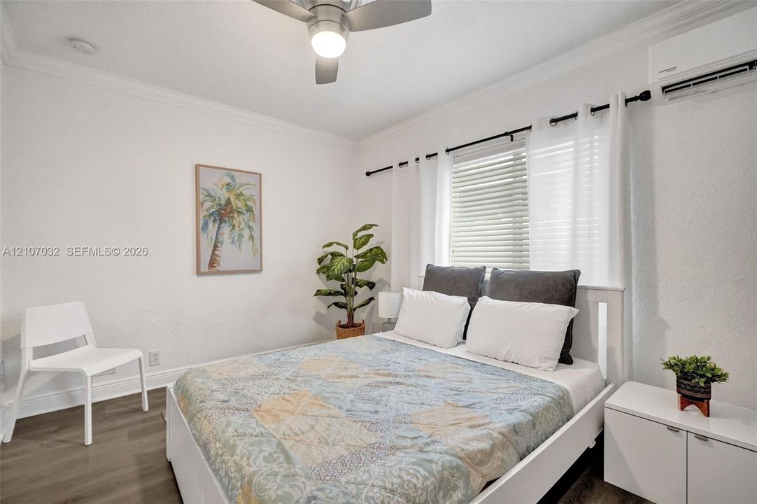 Bedroom featuring wood-finish flooring, crown molding, ceiling fan with integrated lighting, wall-mounted ductless air conditioning unit, and horizontal blinds