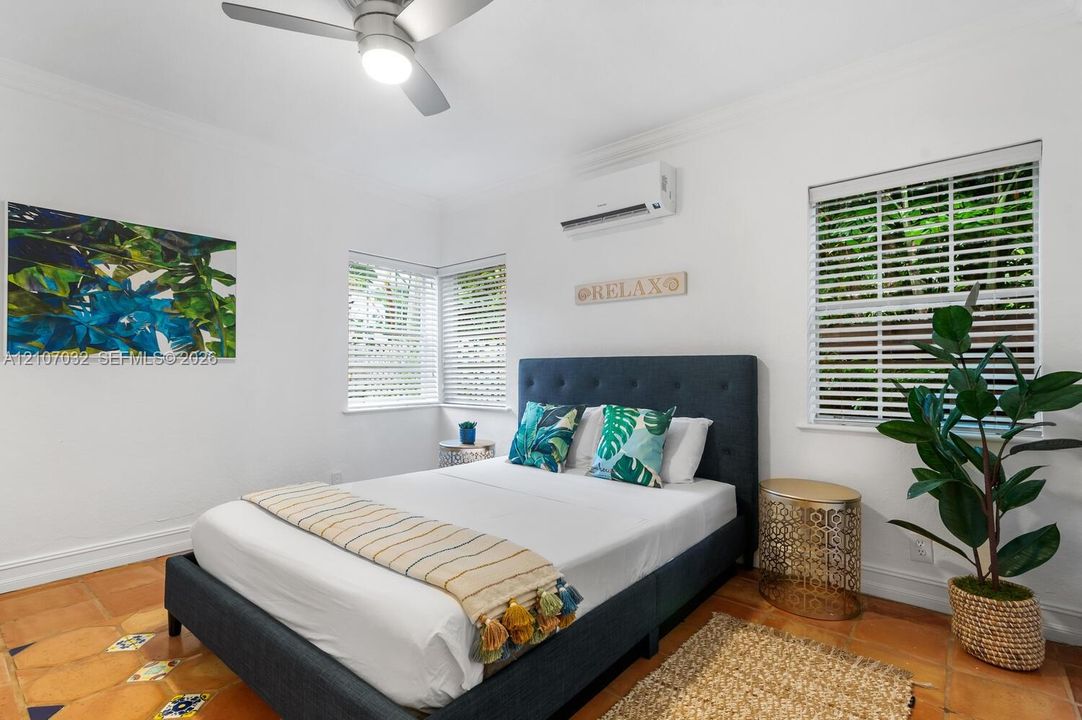 Neutral-toned room featuring terra-cotta tile flooring, a wall-mounted ductless air conditioning unit, a ceiling fan with integrated lighting, and multiple windows with horizontal blinds
