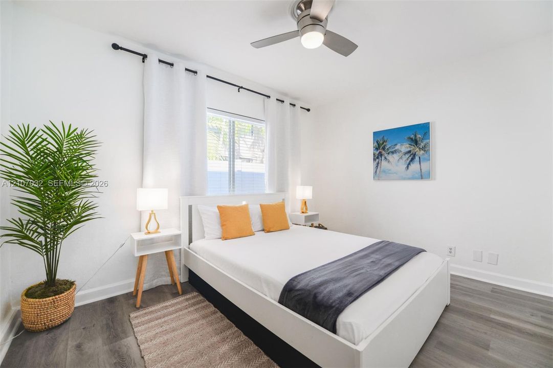 Neutral-toned bedroom featuring wood-finish flooring, a brushed metal ceiling fan with integrated lighting, and a single window framed by an adjustable curtain rod