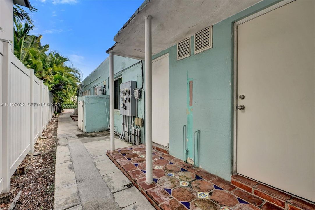 Exterior side entry featuring a covered porch with decorative patterned tile flooring, supported by round structural columns and enclosed by a tall privacy fence