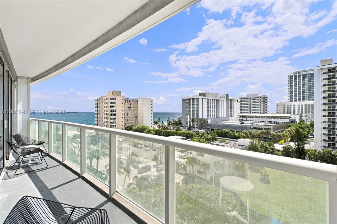 Private covered balcony featuring a glass panel railing, concrete flooring, and panoramic coastal and urban scenery