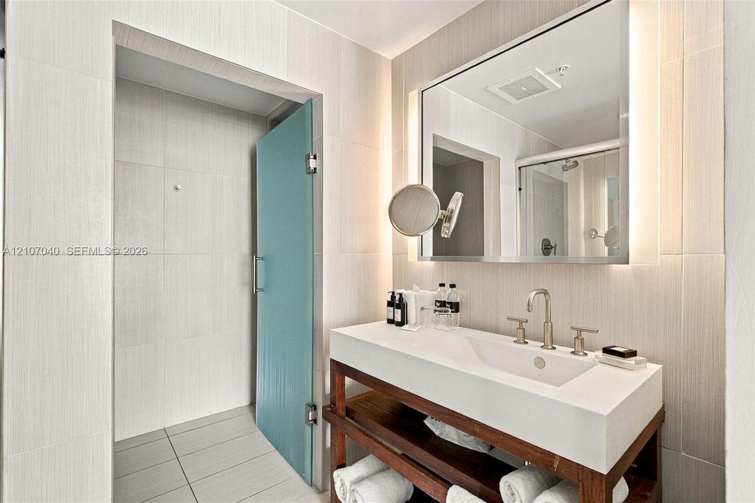 Open-concept vanity featuring a white composite countertop, integrated rectangular sink, metal widespread faucet, wood-finish shelving, and wall-mounted mirror with integrated lighting, finished with floor-to-ceiling tile work and a frosted glass door