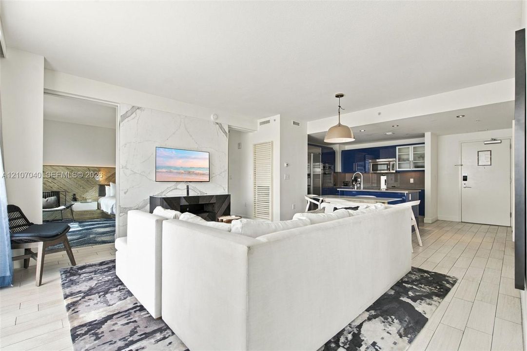 Open-concept living space featuring large-format tile flooring, a floor-to-ceiling stone-surround wall, glossy navy cabinetry, integrated kitchen appliances, and a polished metal pendant light fixture