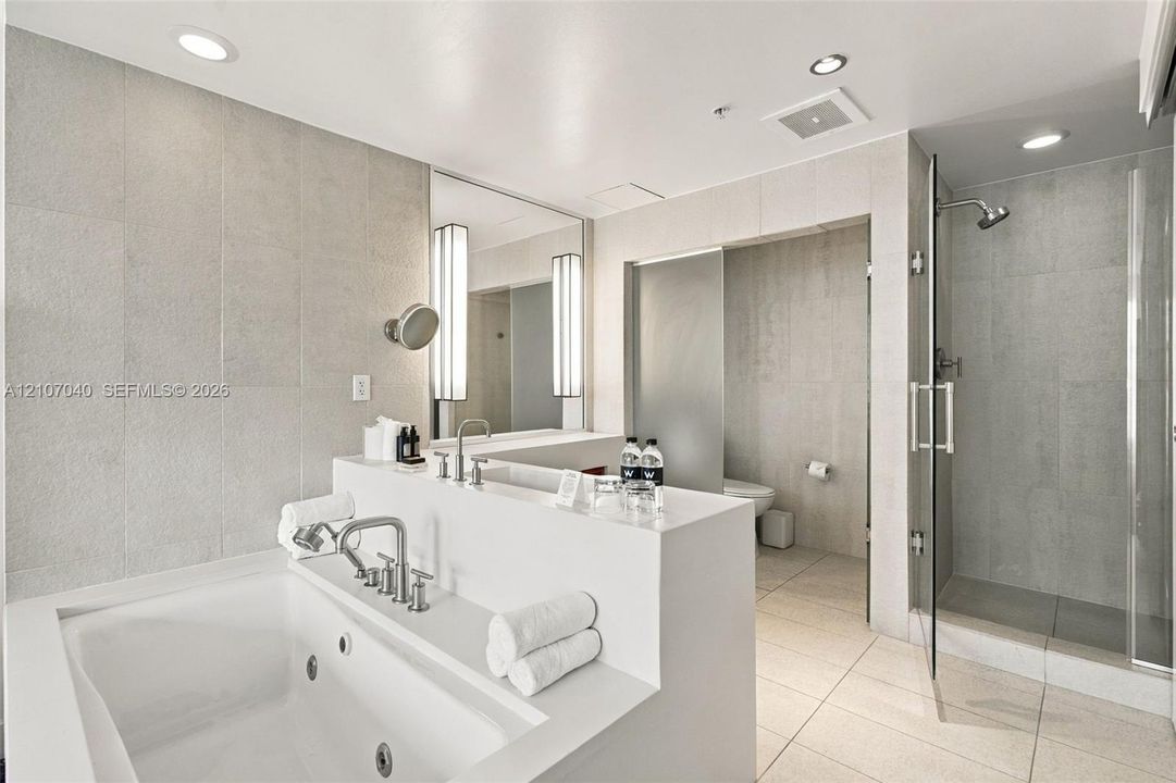 Jetted soaking tub with polished chrome fixtures, walk-in glass-enclosed shower, large-format tile wall cladding, and recessed lighting