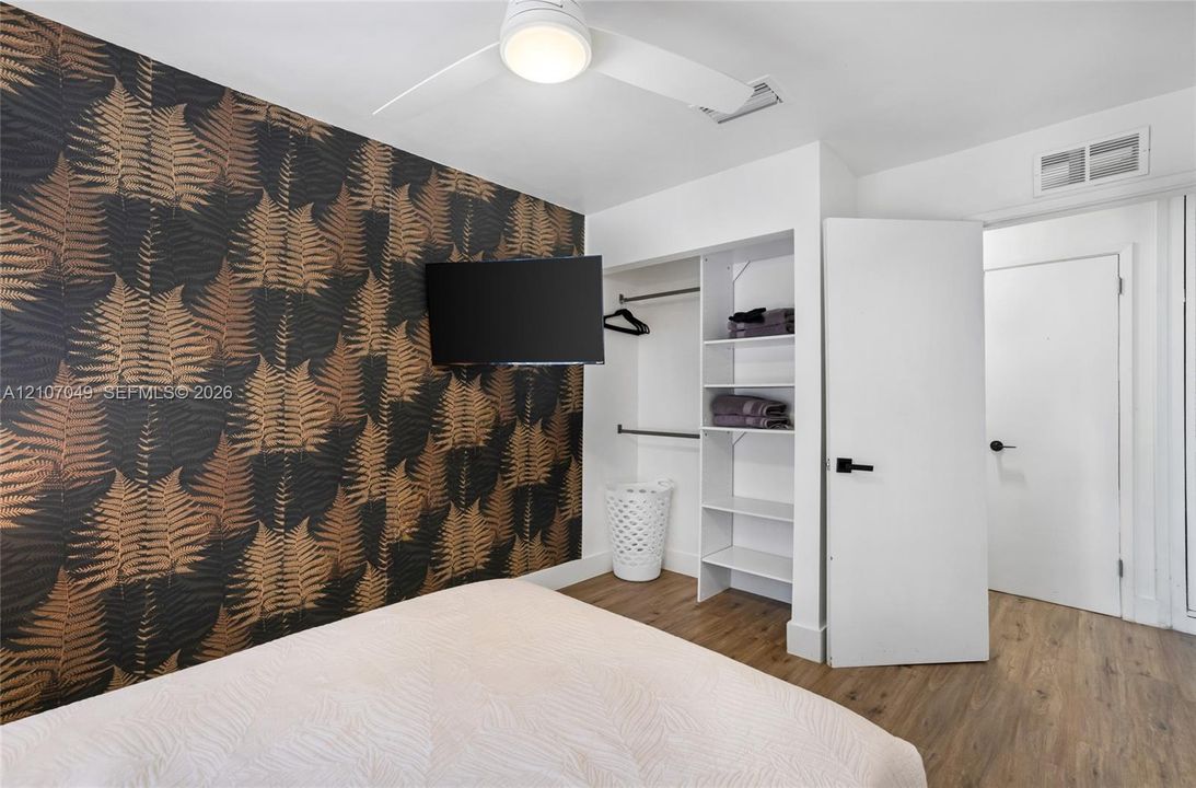 Accent wall with botanical wallpaper, wood-finish flooring, white ceiling fan with integrated lighting, and built-in closet featuring shelving and hanging rods