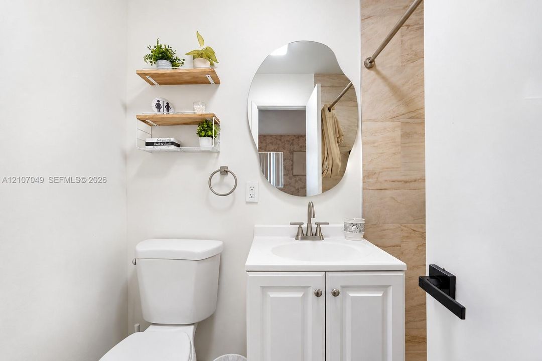 White vanity with a solid surface countertop, organic-shaped wall mirror, two tiered wood-finish wall shelves, and neutral stone-finish tile surround