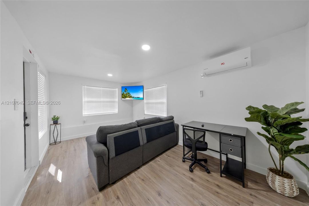Neutral-toned interior space featuring wood-finish flooring, recessed lighting, white walls, horizontal window blinds, and a wall-mounted ductless air conditioning unit