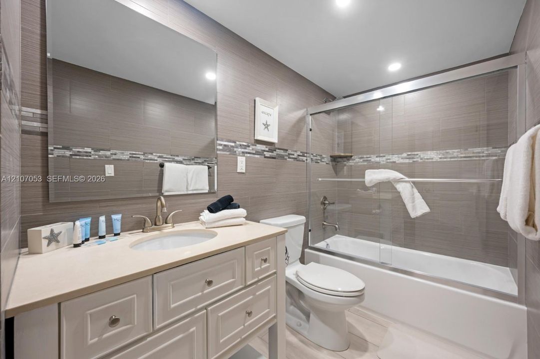 Bathroom featuring a white vanity cabinet with a light-toned countertop, an integrated oval basin, full-width mirror, tile-surround shower-tub combination with sliding glass doors, and horizontal decorative accent tiling