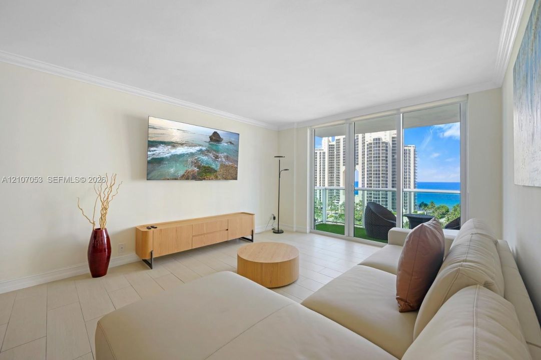 Living area featuring large-format tile flooring, crown molding, and floor-to-ceiling glass sliding doors opening to a balcony with ocean and cityscape scenery