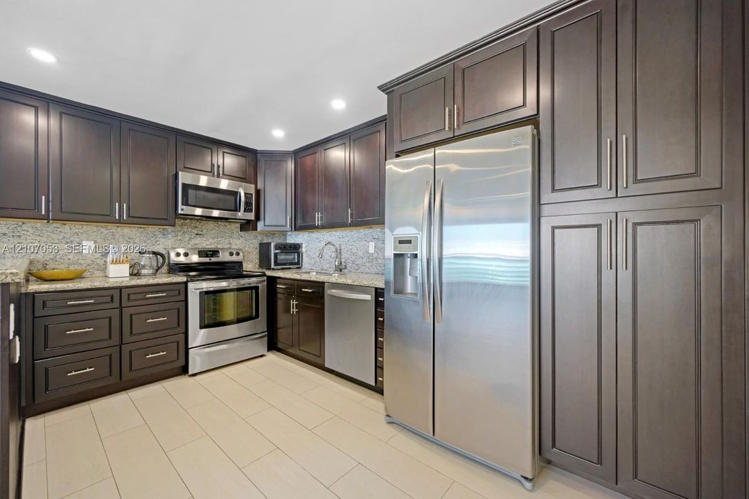 Kitchen featuring dark wood cabinetry, stainless steel appliances, stone-look countertops, tiled backsplash, and recessed lighting