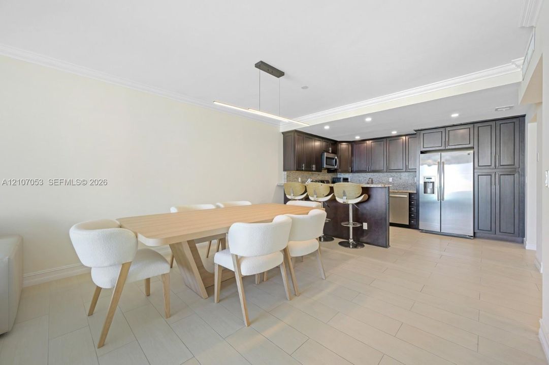 Open-concept kitchen featuring dark wood cabinetry, stainless steel appliances, neutral stone-finish countertops, tile flooring, and a modern linear pendant light fixture