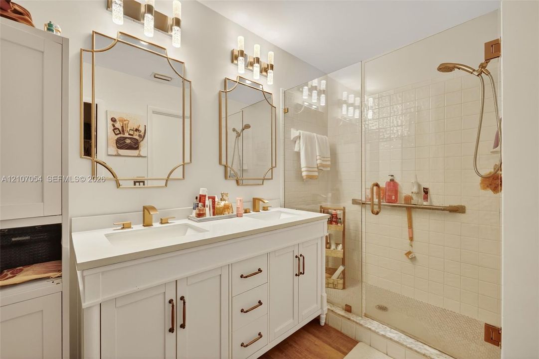 Dual-sink vanity with white cabinetry, quartz countertop, gold-tone hardware, decorative framed mirrors, and glass-enclosed walk-in shower
