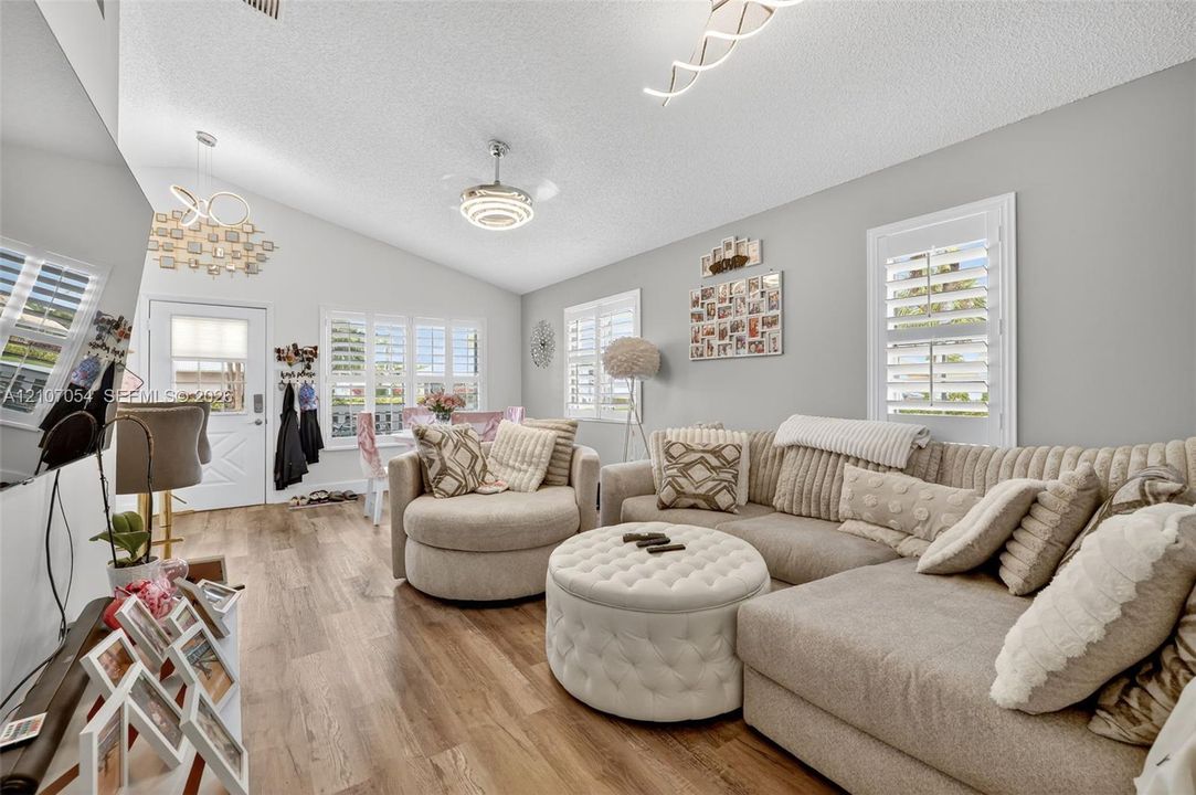 Living area featuring laminate flooring, vaulted ceilings, multi-light chandeliers, and windows outfitted with white plantation shutters
