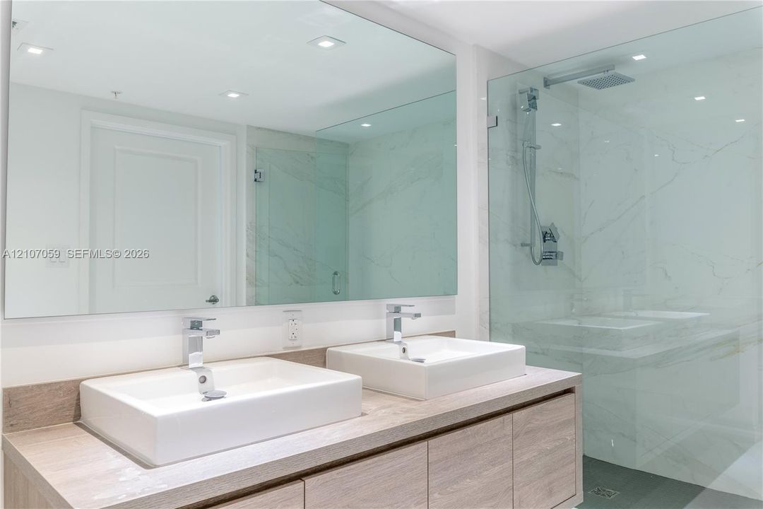 Double vanity featuring wood-finish cabinetry, rectangular vessel sinks, polished chrome faucets, and a frameless glass-enclosed shower with large-format stone-tile surround