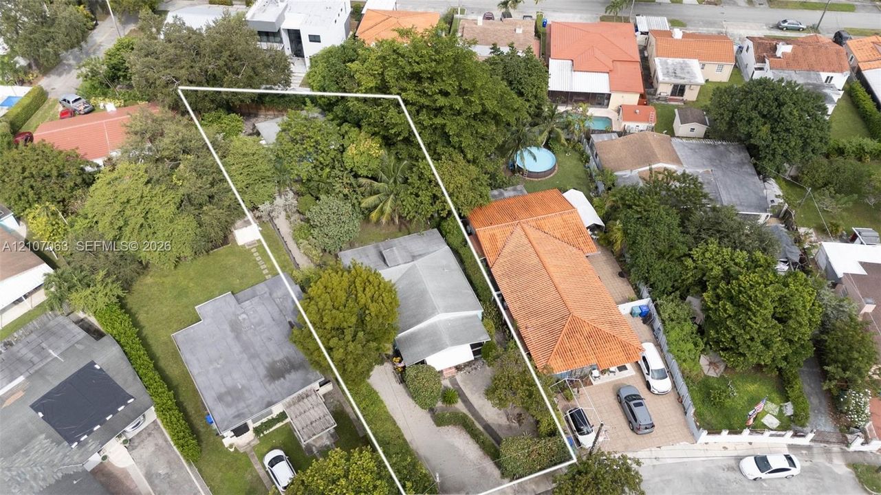 Aerial perspective of residential property featuring a paved driveway, mature trees, manicured landscaping, and a fenced lot