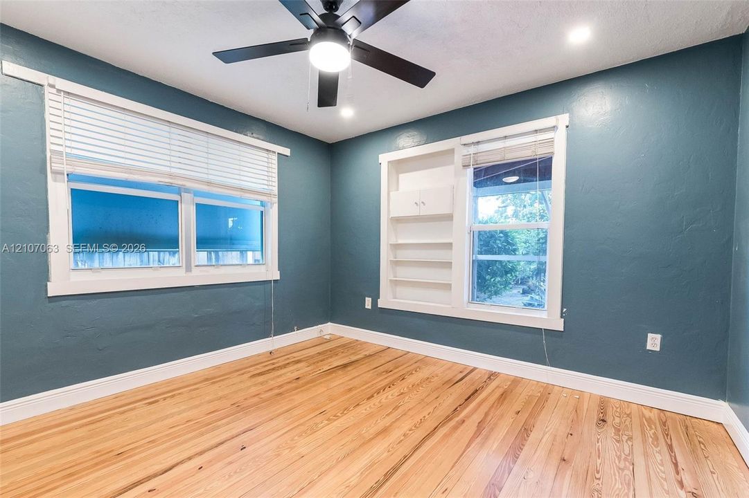 Interior room featuring wood-finish flooring, deep teal wall color, integrated built-in shelving, multi-pane windows with white casing, and a ceiling-mounted light fixture with fan blades
