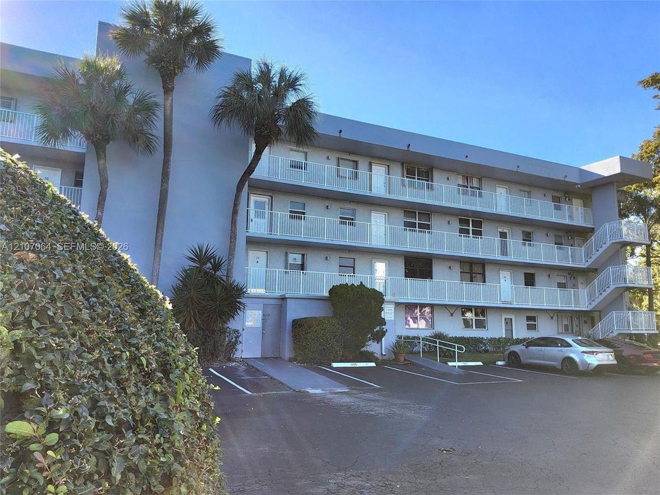 Multi-story building exterior featuring a concrete facade, white metal balcony railings, an external staircase, and mature palm trees