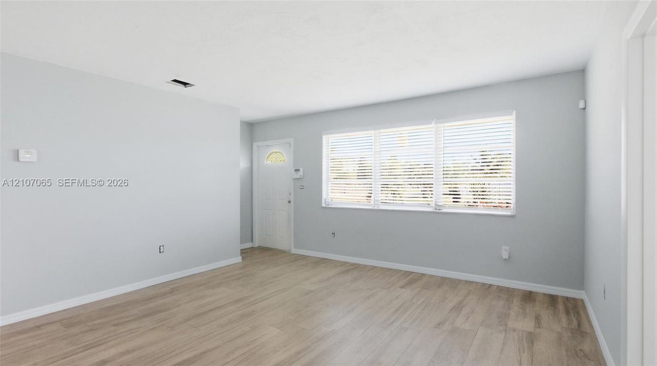 Neutral interior with light-toned wood-finish flooring, white walls, and a large triple-pane window with installed horizontal blinds