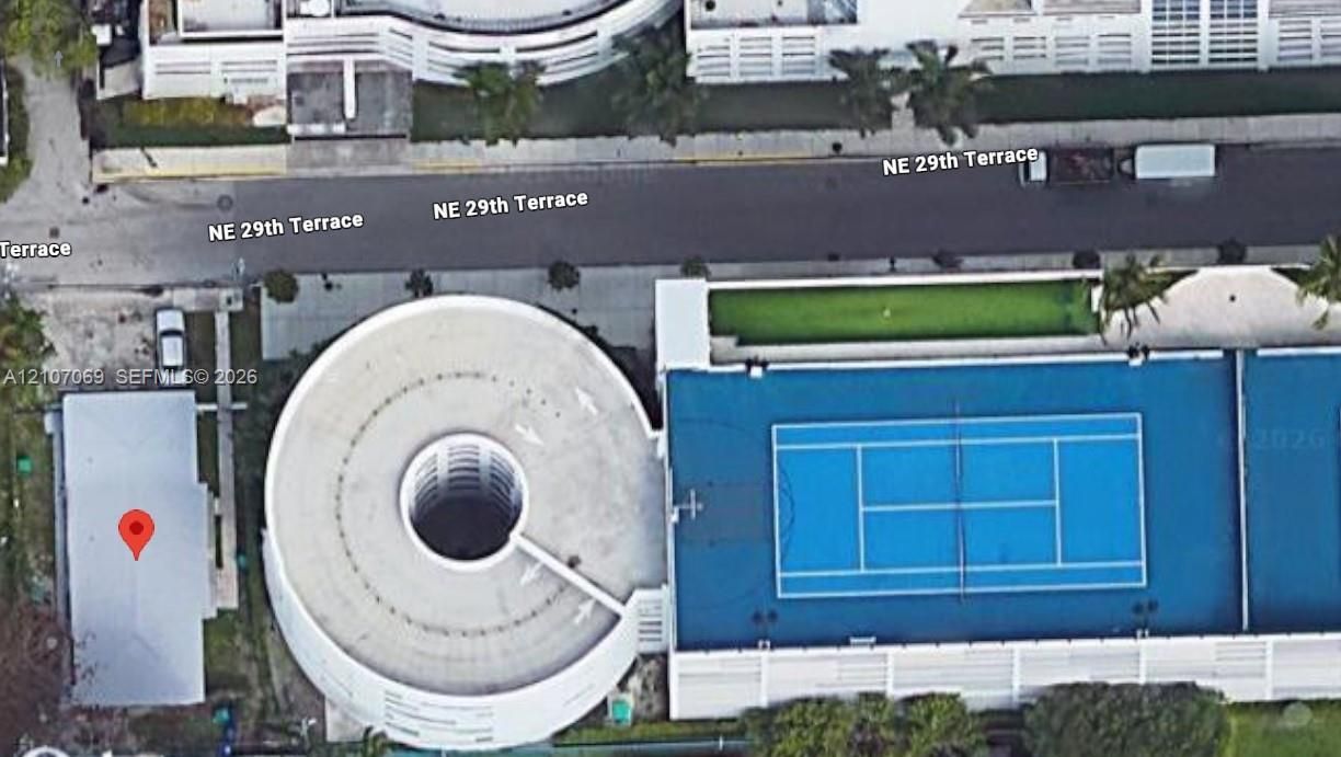 Aerial perspective featuring a hard-surface tennis court, a circular multi-level parking structure, and landscaped green space