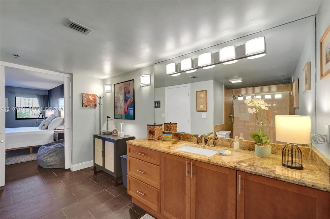 Bathroom vanity with natural stone countertop, integrated rectangular sink, wooden cabinetry, and expansive wall-mounted mirror featuring integrated light fixtures
