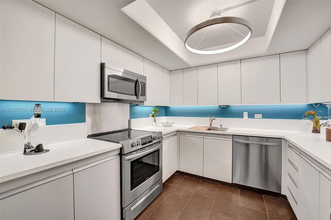 Kitchen featuring white flat-panel cabinetry, solid-surface countertops, stainless steel appliances, tiled flooring, and a recessed ceiling fixture with LED accent lighting