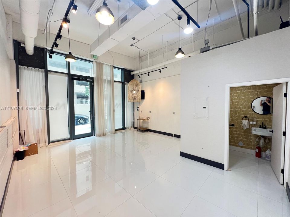 Open-concept interior featuring high ceilings, polished tile flooring, floor-to-ceiling glass paneling, industrial-style track lighting, and a wall-mounted sink with a textured backsplash