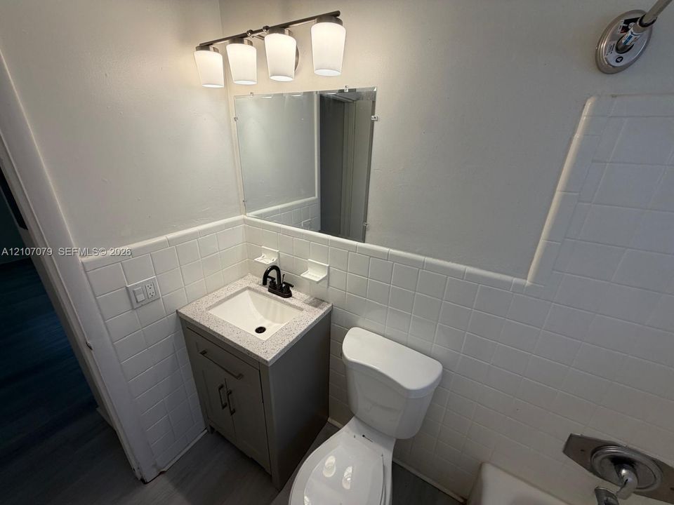Bathroom featuring a vanity cabinet with a stone-composite countertop, undermount sink, matte black faucet, wall-mounted light fixture, and tile wall surfacing