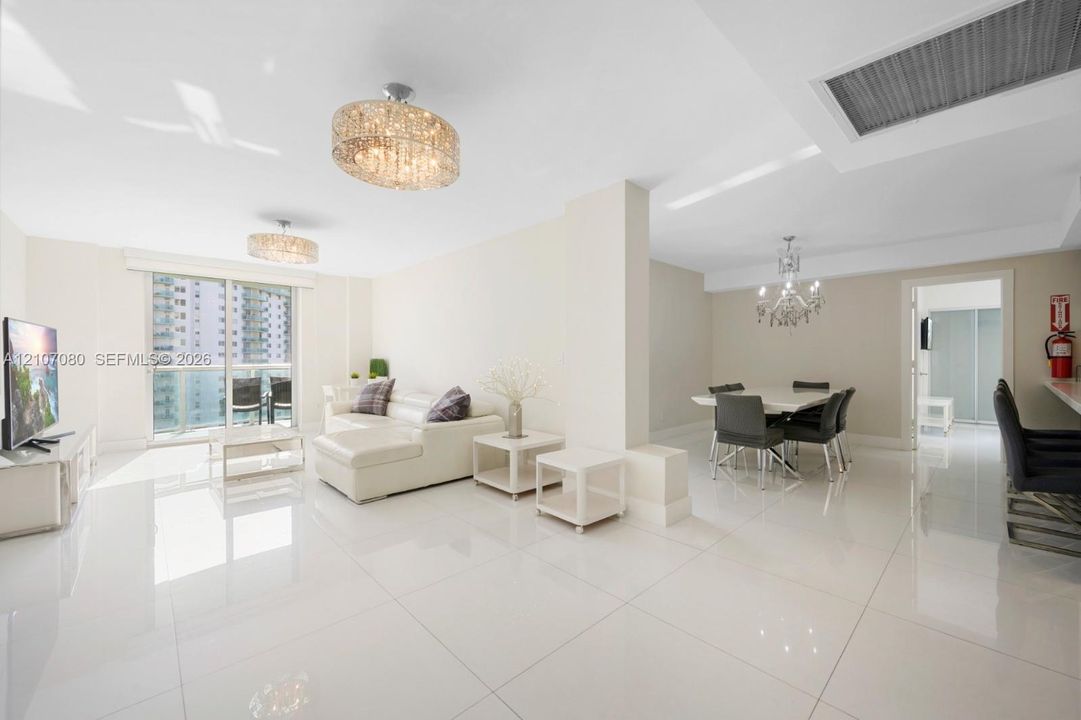 Open-concept living space featuring high-gloss tile flooring, dual contemporary crystal drum chandeliers, a glass-encased crystal dining chandelier, and sliding glass doors leading to a balcony