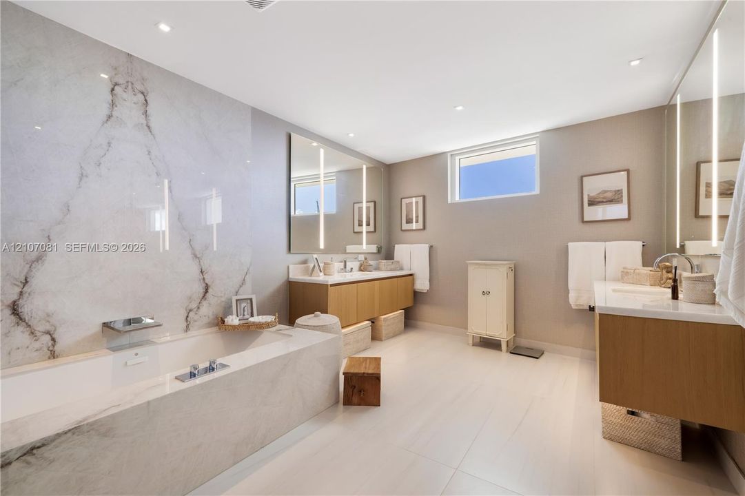 Bathroom featuring a soaking tub with stone-tiled surround, stone slab accent wall, dual floating wood-finish vanities, and vertical integrated mirror lighting