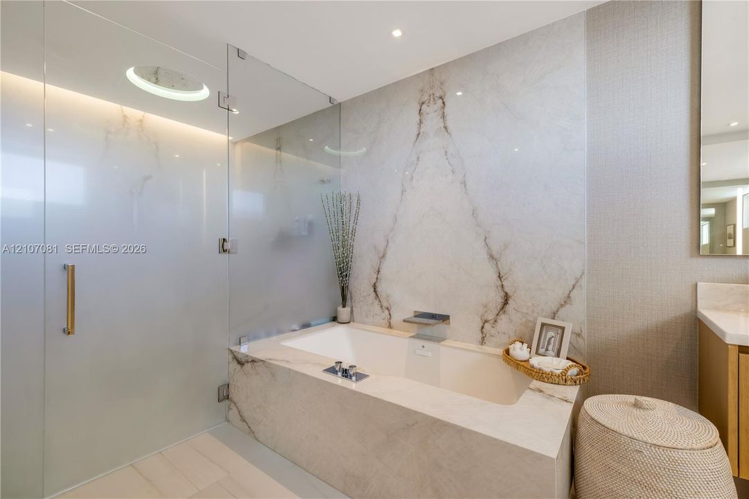 Built-in bathtub featuring full-height natural stone surround, frameless glass shower enclosure, brushed metal fixtures, and recessed ceiling lighting