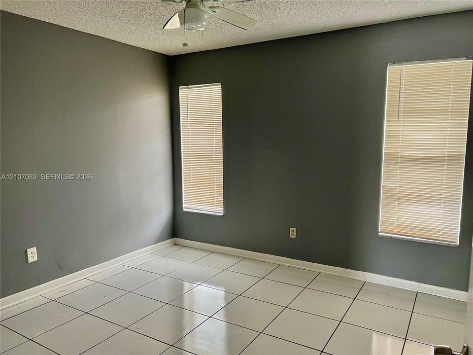 Interior room featuring large-format square tile flooring, neutral grey wall paint, two windows with horizontal blinds, and a ceiling-mounted fan with light fixture