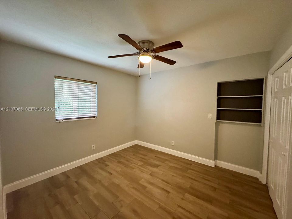 Wood-finish flooring, ceiling fan with integrated lighting, built-in wall shelving, and window with horizontal blinds