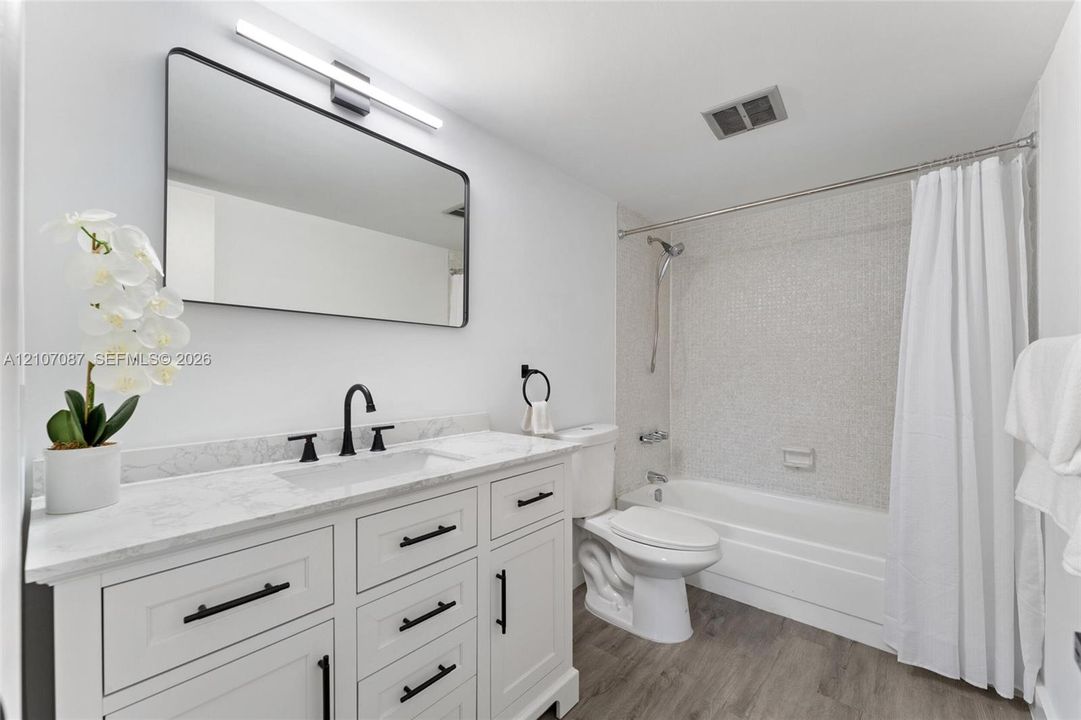 Bathroom featuring a stone-look countertop vanity with matte black hardware, rectangular undermount sink, linear wall-mounted lighting, and a bathtub with tiled surround and wood-finish flooring