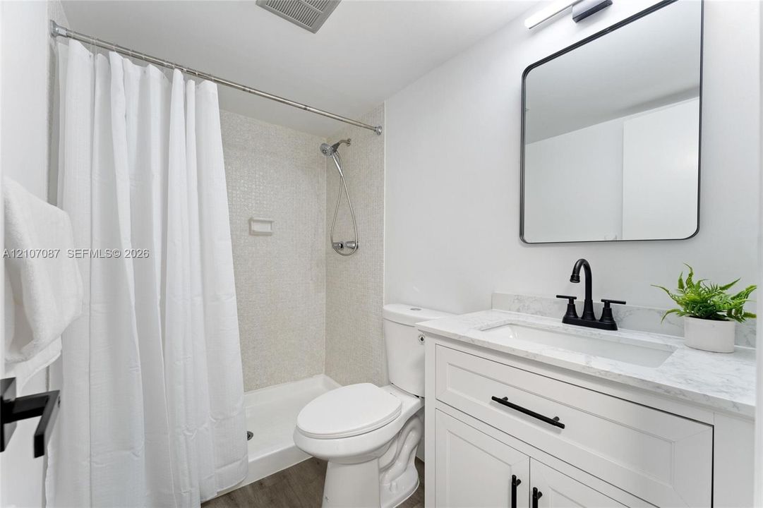 Bathroom featuring a white shaker-style vanity with stone-top surfacing, integrated rectangular sink, matte black hardware, and a shower with mosaic tile surround
