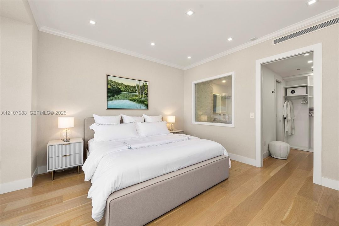 Neutral-toned bedroom featuring light wood-finish flooring, recessed ceiling lighting, crown molding, and an interior window to bathroom with switchable privacy glass