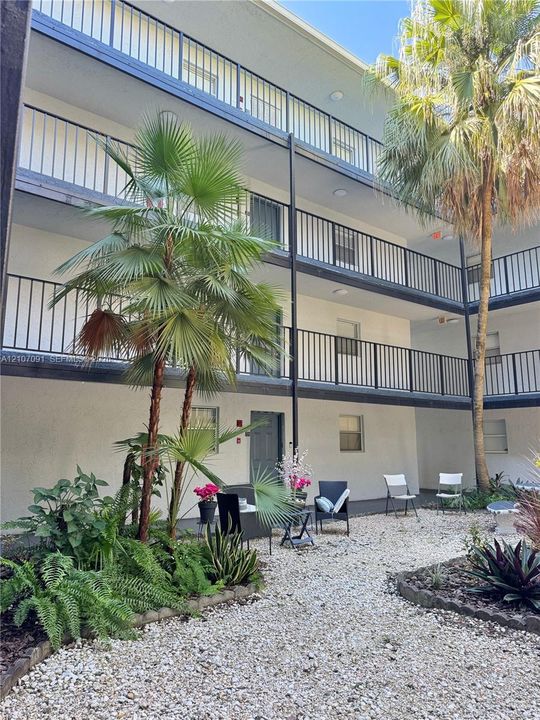 Multi-story exterior featuring metal railed balconies, neutral stucco finish, and landscaped gravel courtyard with mature palms
