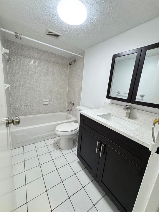Bathroom featuring a dark-finish vanity with a light-toned stone countertop, integrated sink, tiled shower surround with mosaic detailing, and white floor tiling