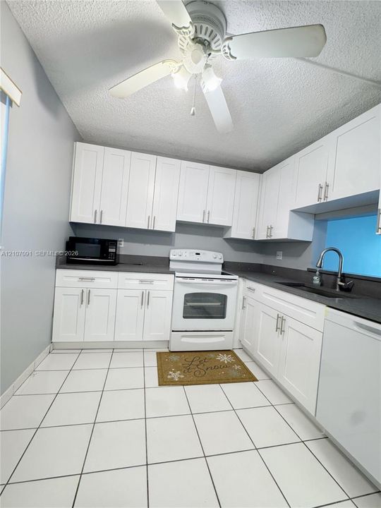 Kitchen featuring white shaker-style cabinetry, dark stone-finish countertops, ceramic tile flooring, a dual-handle gooseneck faucet, and a ceiling-mounted light fixture with an integrated fan