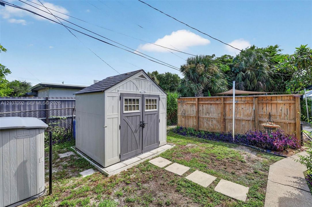 Exterior storage shed featuring dual entry doors, shingle-style roof, and decorative window panes, complemented by a wood fence perimeter and stone paver walkway