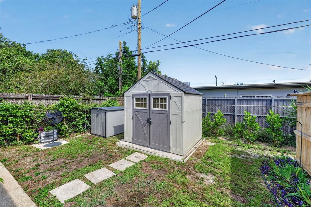 Exterior storage structure featuring a gable roof, double doors with glass paneling, and a reinforced foundation pad, situated within a fenced backyard