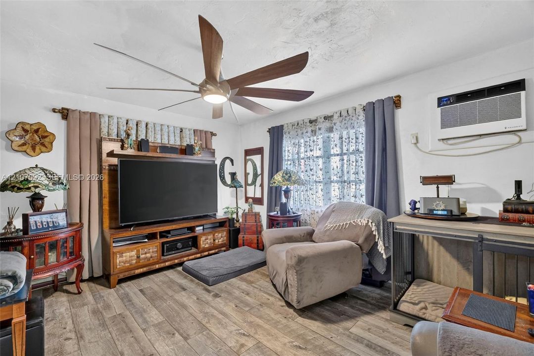 Living area featuring wood-finish flooring, a multi-blade ceiling fan with integrated lighting, and a wall-mounted air conditioning unit