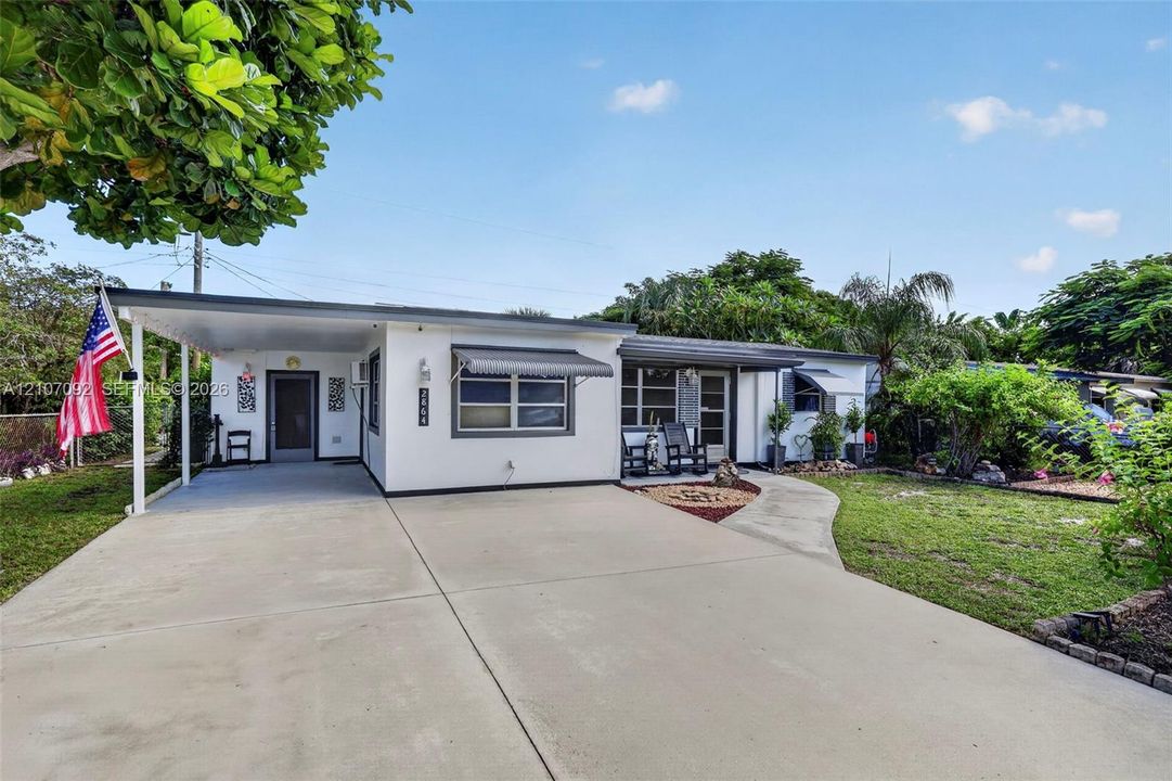 Single-story exterior featuring a concrete driveway, attached carport, metal window awnings, and a covered front entryway