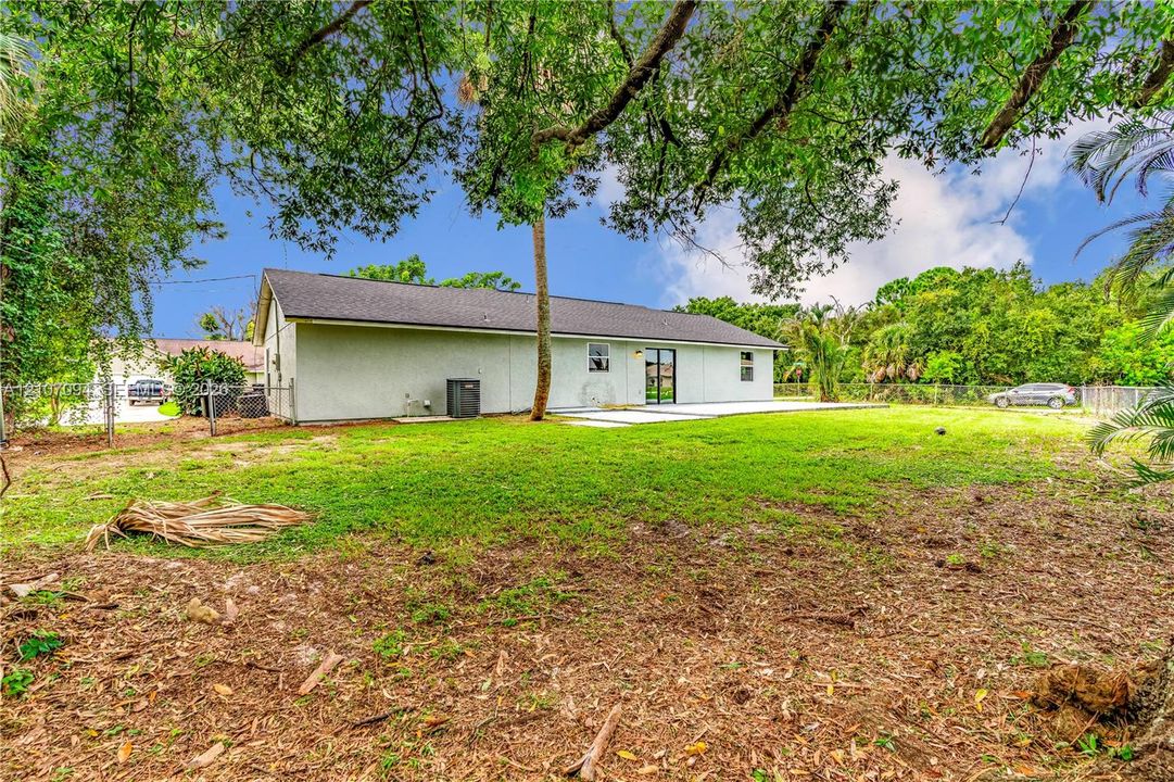 Backyard featuring a concrete patio, light-colored stucco exterior, shingle roof, chain-link perimeter fencing, and mature trees