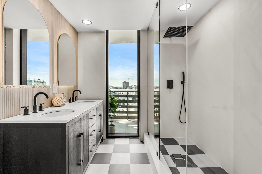 Double vanity with a white solid-surface countertop, dark cabinetry, arched wall mirrors, matte black fixtures, and a glass-enclosed shower featuring a rain-style showerhead and checkerboard tile flooring